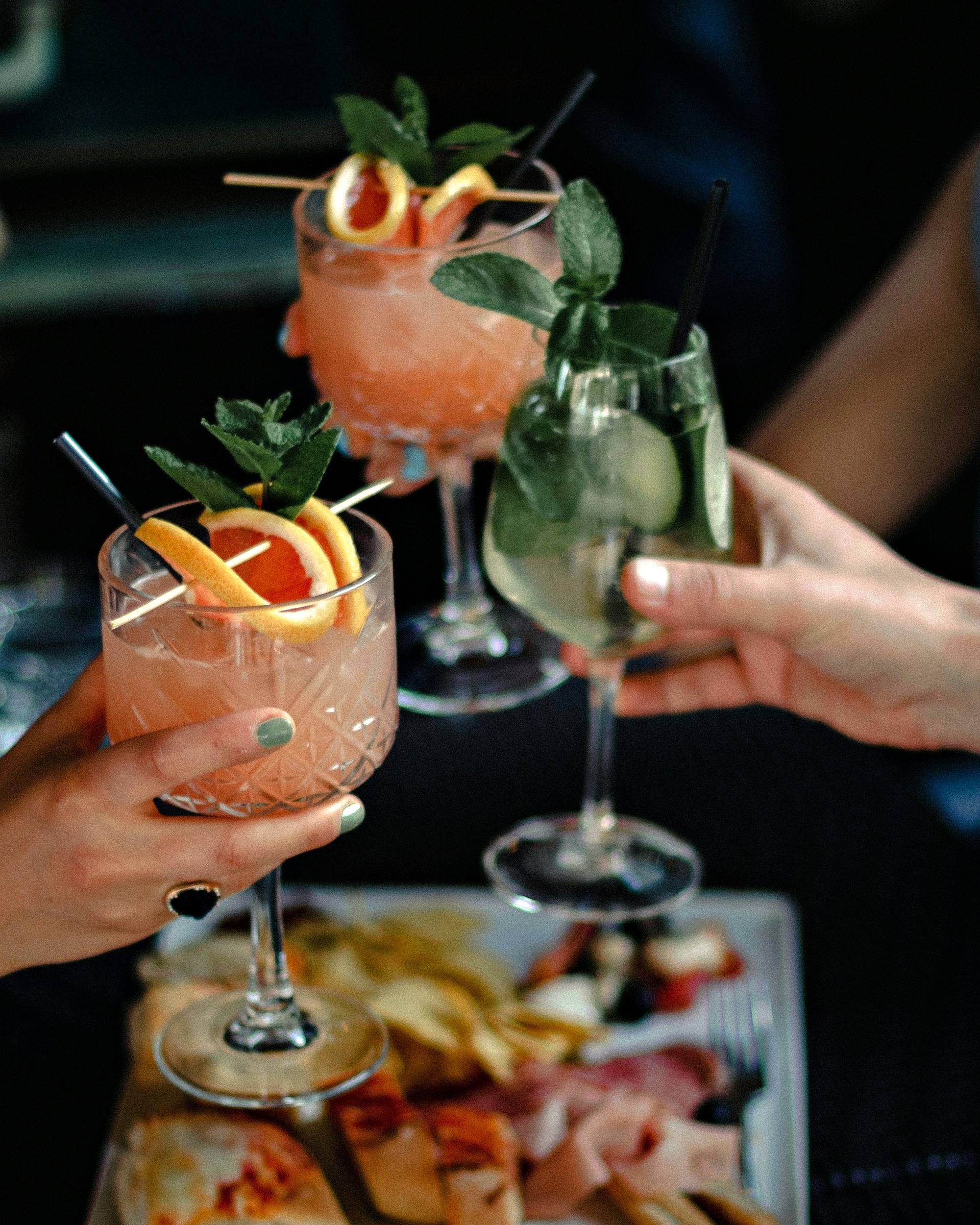 Drei Hände stoßen mit Spritz-Gläsern an, darunter ein Brett mit Prosciutto, Brot und Chips