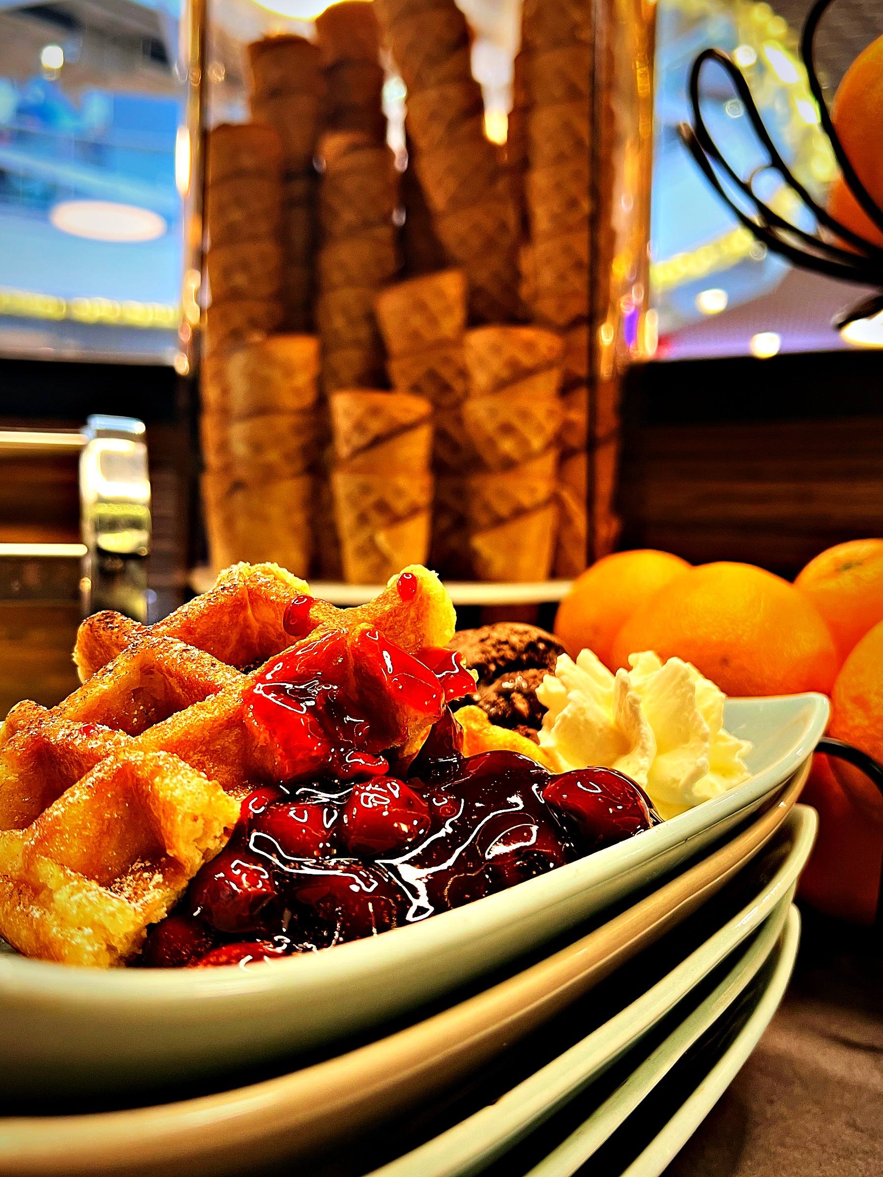 Belgische Waffel mit Kirschsauce, einer Kugel Eis und Schlagobers, dahinter Orangen